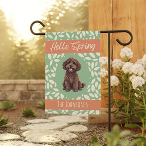May include: A garden flag with a brown dog illustration, the text "Hello Spring" and "The Johnson's" on a green and peach background. The flag is displayed on a black metal stand, surrounded by greenery and a stone pathway.