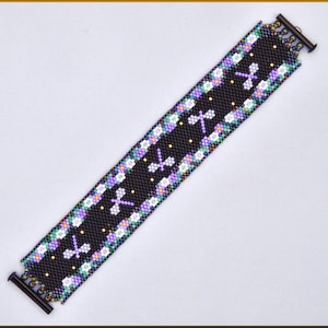 Pode incluir: Uma pulseira larga de missangas com fundo preto e um padrão repetitivo de libelinhas em roxo claro. As bordas são decoradas com um mosaico colorido de missangas brancas, roxas e verdes. A pulseira é fixada com barras de metal escuro.