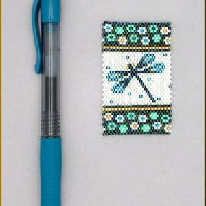 Puede incluir: Un marcapáginas de cuentas con fondo blanco y un diseño de libélula azul. El marcapáginas tiene ribetes verdes y dorados.