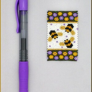 May include: A purple pen with a black ink cartridge is next to a beaded bracelet with a white background and a pattern of bees and flowers. The bracelet has a green border with purple and yellow flowers.