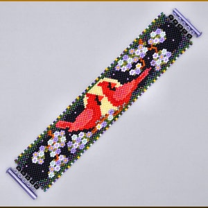May include: A wide beaded cuff bracelet featuring a black background with two red cardinal birds perched on branches with white and purple blossoms. The bracelet is finished with a metallic purple slide clasp at both ends, securing the woven beadwork.