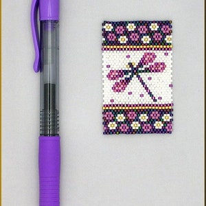 May include: A beaded dragonfly charm with a white background and purple, pink, and white floral borders.