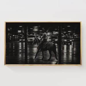 Könnte beinhalten: Ein schwarz-weiß gerahmter Wandkunstdruck, der eine Person mit Cowboyhut zeigt, die auf einem Pferd durch eine dunkle, nasse Stadtstraße bei Nacht reitet. Die Szene zeigt verschwommene Autoscheinwerfer und Lichtreflexionen auf dem Boden.