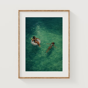 Puede incluir: Impresión enmarcada que muestra a dos personas en agua turquesa. Una está en un flotador inflable con estampado de cebra, sosteniendo una bebida. La otra persona está flotando. La impresión está en un marco de madera.