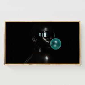 Puede incluir: Una impresión de arte mural enmarcada que muestra a una persona de piel oscura con gafas de sol de visera reflectante y soplando una burbuja grande, redonda y de color verde azulado sobre un fondo negro sólido de alto contraste.