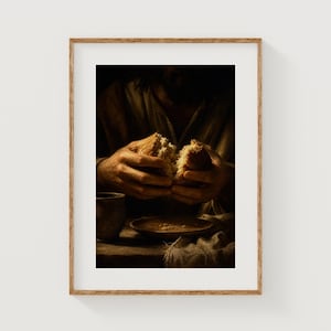 May include: A framed print depicts a person breaking bread. The image features a dark, warm color palette with a focus on the hands and the bread. A cup and a plate with food are also visible, suggesting a religious or historical theme.