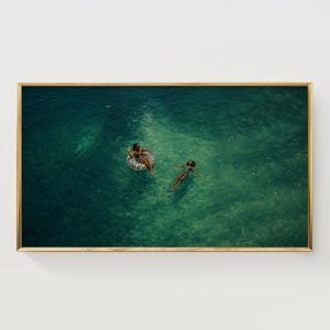 Puede incluir: Impresión enmarcada que muestra a dos personas en agua turquesa. Una persona flota en un flotador inflable con estampado de cebra, la otra flota cerca. El agua clara y la escena se capturan desde arriba.