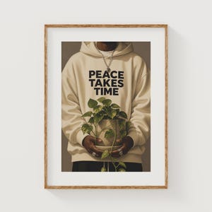 Pode incluir: Impressão emoldurada com uma pessoa vestindo um moletom creme com a frase "PEACE TAKES TIME" em preto. A pessoa está segurando uma planta em vaso com folhas verdes. A impressão está em uma moldura de madeira.