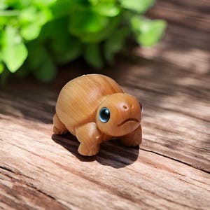 Puede incluir: Una pequeña figura de tortuga de madera con ojos azules. La tortuga está pintada de color marrón claro y tiene una textura detallada.