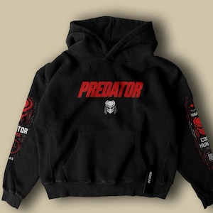 Può includere: Felpa con cappuccio nera con la scritta "PREDATOR" in rosso e una grafica della maschera Predator bianca. Le maniche presentano disegni grafici e testo rossi e bianchi. È attaccata un'etichetta nera con "DESIGN".