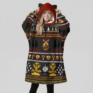 May include: A black hooded blanket with a red hood and cat ears. The blanket features a pattern with Egyptian symbols, including cobras, scarabs, and hieroglyphs. The word "SCARAB" is visible on the front.