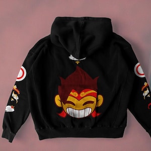 Puede incluir: Sudadera con capucha negra con un personaje de dibujos animados en la espalda. El personaje tiene el pelo rojo, una diadema amarilla y una gran sonrisa. Las mangas tienen pequeñas imágenes de dibujos animados de nubes y pájaros.