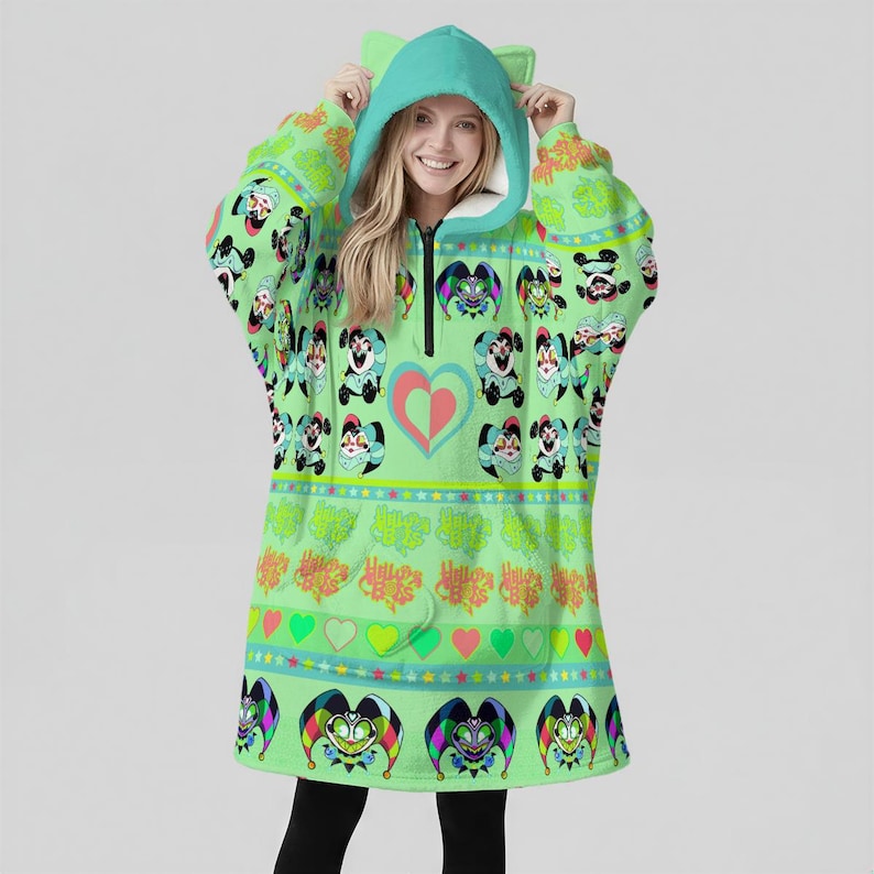 May include: A light green hooded blanket with a zip-up front and cat ears on the hood. The blanket features a repeating pattern of cartoon characters, hearts, and jester faces. The blanket is worn over black leggings.