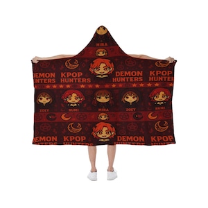 May include: A hooded blanket with a repeating pattern of cartoon characters, moons, stars, and the text "DEMON KPOP HUNTERS" in red and white on a dark red background. The blanket is being held up by a person.