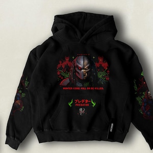 Può includere: Felpa con cappuccio nera con una stampa grafica di un personaggio di Predator. La stampa presenta il viso del Predator, uno sfondo di giungla e il testo "HUNTER GOOD. KILL OR BE KILLED." e "プレデター PREDATOR".