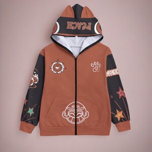 Pu&ograve; includere: Una felpa con cappuccio marrone con zip, con un design in bianco e nero che presenta un viso di scimmia, stelle e il testo "GOOD MONK" e "PEACH".