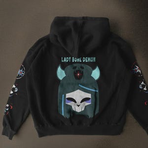 May include: Black hooded sweatshirt with the text "LADY BONE DEMON" above a graphic of a skull-like figure with teal horns and hair. The sleeves feature compass and bone designs.
