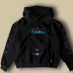 May include: Black hoodie with the text "HELLUVA BOSS FIZZAROLLI" in teal and pink. The hoodie features a front pocket and decorative designs on the sleeves. A heart and teal design is on the front.