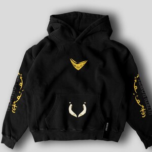 May include: Black hooded sweatshirt with a kangaroo pocket and a drawstring hood. The sweatshirt features gold-colored graphics on the chest, sleeves, and pocket. The sleeves have text in a stylized font. A small black tag with the word "DESIGN" is on the lower right.
