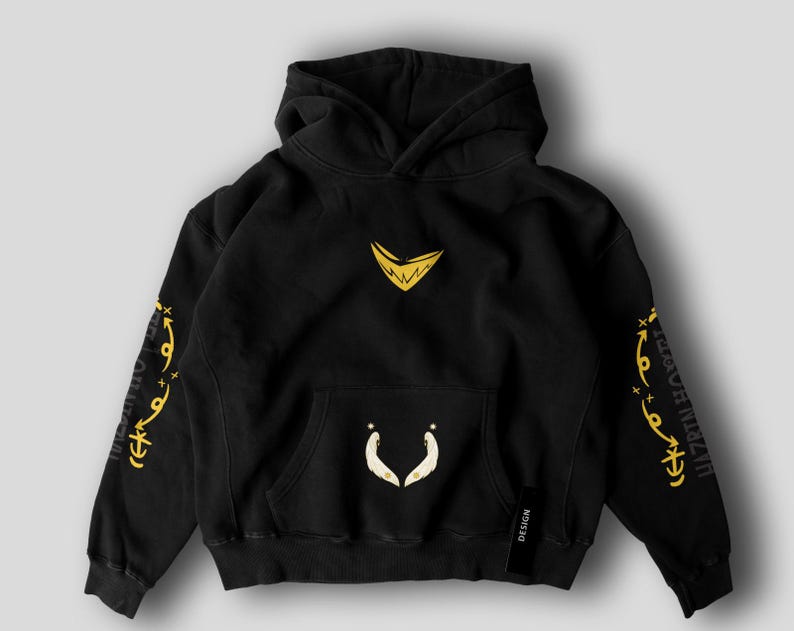 May include: Black hooded sweatshirt with a kangaroo pocket and a drawstring hood. The sweatshirt features gold-colored graphics on the chest, sleeves, and pocket. The sleeves have text in a stylized font. A small black tag with the word "DESIGN" is on the lower right.