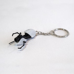 May include: A miniature portal gun keychain, predominantly black and white, with a silver chain and keyring. The design is based on a video game. The keychain is a small, detailed replica.