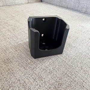 Pu&ograve; includere: Un supporto in plastica nera, angolare, con un'apertura rettangolare e diversi fori. Il supporto &egrave; progettato per essere montato su una parete o altra superficie. L'oggetto &egrave; appoggiato su una superficie strutturata di colore neutro.