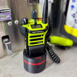 May include: A neon green and black Motorola two-way radio in a black charging base with a red stripe. The radio has the text "MOTOROLA" and "APX 6000" printed on it. The base has the text "FCDF" printed on it.