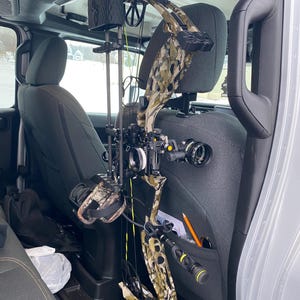 May include: A compound bow with a camouflage pattern is secured inside a vehicle. The bow features a sight, stabilizer, and other accessories. The bow is positioned near the seat, with additional gear visible in the background.