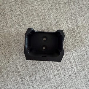 Pu&ograve; includere: Un oggetto nero rettangolare stampato in 3D con due fori circolari. L'oggetto ha una superficie strutturata e una forma unica, forse un supporto o una base per un altro oggetto. L'oggetto &egrave; su una superficie grigia.