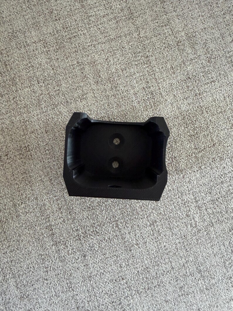 Pu&ograve; includere: Un oggetto nero rettangolare stampato in 3D con due fori circolari. L'oggetto ha una superficie strutturata e una forma unica, forse un supporto o una base per un altro oggetto. L'oggetto &egrave; su una superficie grigia.