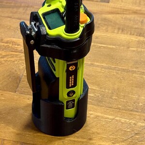 Könnte beinhalten: Ein Funkgerät in einer schwarzen Ladeschale. Das Funkgerät ist limettengrün und schwarz, mit einem kleinen Bildschirm und einer Antenne. Die Halterung ist schwarz und steht auf einer Holzoberfläche. Das Funkgerät hat einen Clip an der Seite.