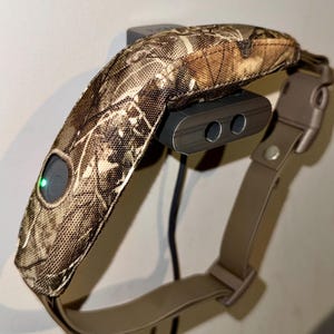 May include: A dog training collar with a camouflage pattern. The collar is tan and brown with a black charging station. The collar has a green light and a buckle closure.