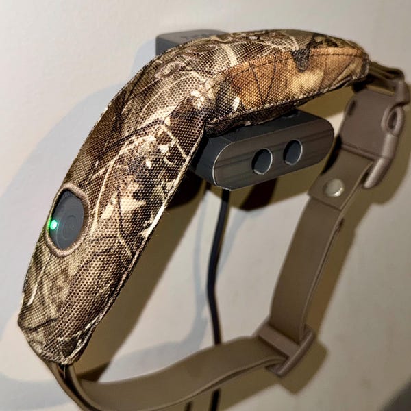 Może przedstawiać: Obroża treningowa dla psa z kamuflażem. Obroża jest brązowo-beżowa z czarną stacją ładującą. Obroża ma zielone światło i zapięcie na klamrę.