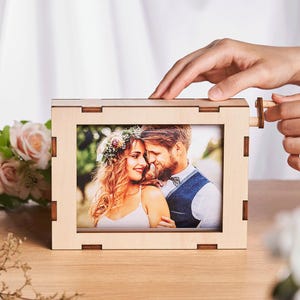 May include: A light-coloured wooden photo frame with a picture of a couple. The frame has a unique design with interlocking joints and a small wooden peg for opening. The photo shows a smiling couple, likely a wedding photo.