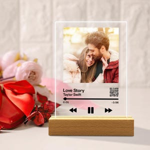 Puede incluir: Una pantalla fotográfica de acrílico transparente con una base de madera. La pantalla presenta una foto de una pareja y los detalles de la canción "Love Story" de Taylor Swift. El diseño incluye una barra de reproducción y un código QR.