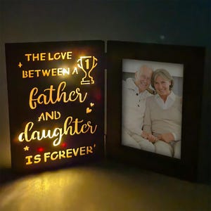 May include: A black wooden photo frame with a lit-up panel. The panel reads "THE LOVE BETWEEN A father AND daughter IS FOREVER" in gold script, with a trophy graphic. The right side holds a photo of an older couple.