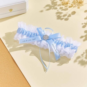 May include: A white and blue garter with a satin ribbon and a rhinestone embellishment. The ribbon is personalized with the text "Mr. & Mrs. Smith 8th June 2024".