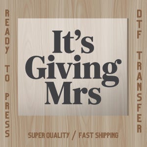 May include: A rectangular DTF transfer with the text "It's Giving Mrs" in a bold, dark gray font. The words are centered on a light beige background, with the words "Ready to Press" and "DTF Transfer" on the sides.
