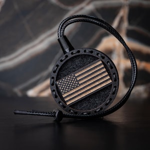 Puede incluir: Un colgante circular negro con una incrustación de madera de la bandera estadounidense. La bandera presenta un diseño detallado con estrellas y rayas. El colgante está unido a un cordón de cuero negro, creando un accesorio patriótico.