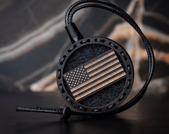 Puede incluir: Un colgante circular negro con una incrustación de madera de la bandera estadounidense. La bandera presenta un diseño detallado con estrellas y rayas. El colgante está unido a un cordón de cuero negro, creando un accesorio patriótico.