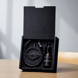 Puede incluir: Caja de madera negra abierta, que contiene un collar con un colgante circular con el logotipo "MINI" y una pequeña botella etiquetada "PURE LEATHER". La caja está abierta, mostrando el contenido.