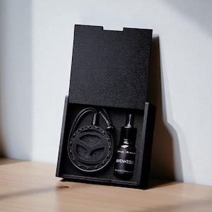 May include: A black wooden box with a pendant necklace and a small bottle of liquid. The pendant has a circular design with a stylized bird emblem. The bottle is labeled "AVEN-TOS".