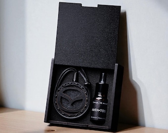 Puede incluir: Una caja de madera negra con un collar con colgante y un pequeño frasco. El colgante tiene un diseño circular con un emblema de pájaro estilizado. El frasco está etiquetado como "AVEN-TOS".