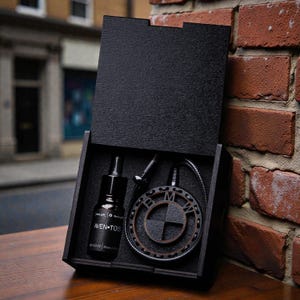 Może przedstawiać: Otwarta czarna drewniana skrzynka, zawierająca małą butelkę i wisiorek z logo BMW. Wisiorek jest okrągły z ciemnym wykończeniem. Na butelce widnieje biały napis "AVEN-TOS". Skrzynka stoi na tle ceglanej ściany.