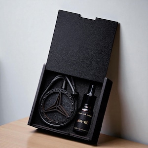 Puede incluir: Una caja de madera negra con la tapa abierta, que revela un collar con un colgante en forma de estrella y una botella pequeña. El colgante tiene un marco circular y un diseño de estrella de tres puntas. La botella tiene una etiqueta negra con texto dorado "KIR-KE".