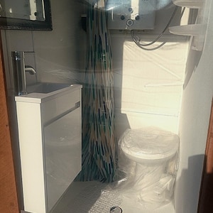 Puede incluir: Un pequeño baño blanco con un lavabo blanco, un inodoro blanco y una ducha con una cortina de ducha estampada. La ducha tiene un calentador de agua blanco con el texto "16L" en él.