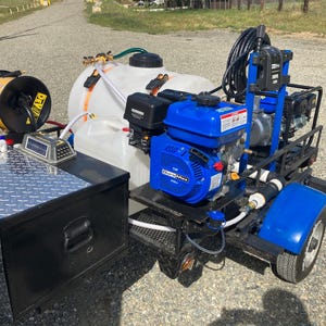 May include: A blue and black gas-powered pressure washer with a white tank, a hose reel, and a black toolbox. The pressure washer is mounted on a small trailer with two wheels.