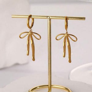 May include: A pair of gold hoop earrings with a dangling bow design. The earrings are displayed on a gold metal stand.