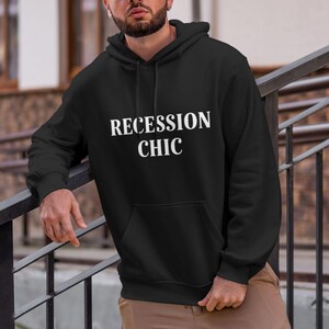 May include: Black hooded sweatshirt with the text "RECESSION CHIC" in white block letters. The sweatshirt has a front pocket and a drawstring hood. The model is wearing khaki pants.