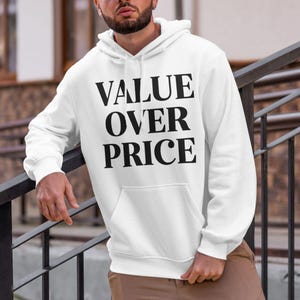 May include: White hoodie with the text "VALUE OVER PRICE" in large, bold, black letters. The hoodie has a front pocket and a hood. The person is wearing brown pants and is leaning against a black metal railing.
